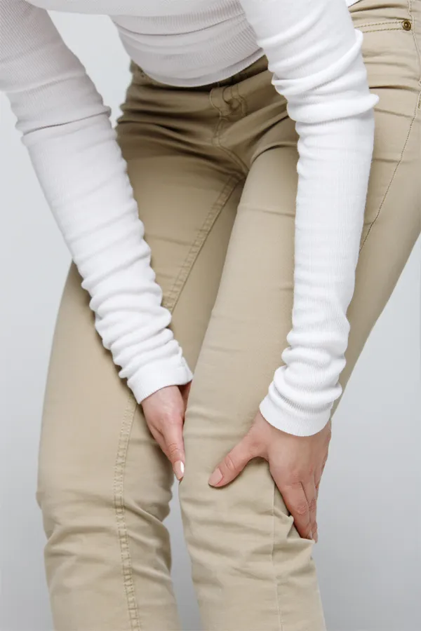 Arthritis-Treatment A woman wearing a white long-sleeve shirt and tan pants, clutching her knee, suffering from arthritis. Get arthritis treatment from ProCare Wellness Institute in Savannah.