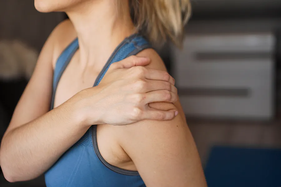 Frozen-Shoulder-Clinic A close-up view of a woman in a blue tank top, gripping her own shoulder with a look of discomfort. Receive expert Frozen Shoulder treatment by ProCare Wellness Institute located in Savannah.
