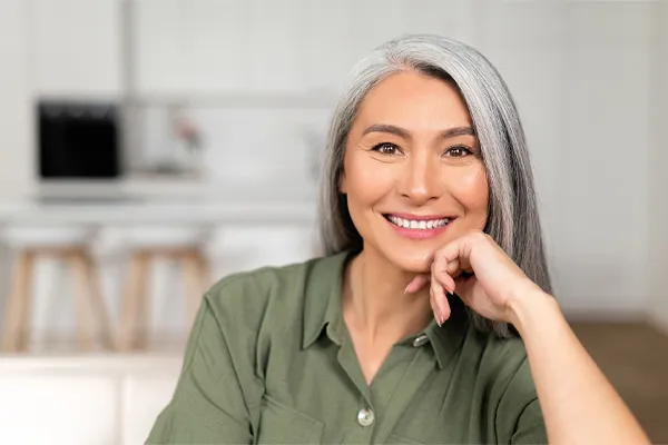 An asian woman with long gray hair and a green shirt sitting and smiling on her couch, happy with her menopause care from [NAME} in Savannah.