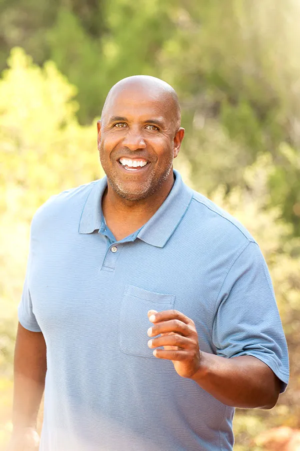 Non-Invasive-Wounds-Treatment Middle-aged African American man smiling broadly, dressed in a casual blue polo shirt. He is outdoors walking on a sunlit trail happy after receiving advanced Non-Healing Wound Care from ProCare Wellness Institute in Savannah.