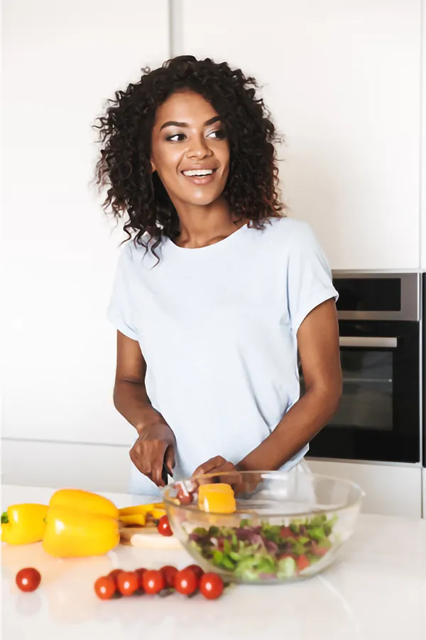 Tendonitis-Treatment A joyful African American woman preparing food in a modern kitchen. She is chopping yellow bell peppers and has a variety of fresh vegetables like tomatoes and lettuce on the counter, assembling a salad after receiving advanced Non-Healing Wound Treatment by ProCare Wellness Institute in Savannah.